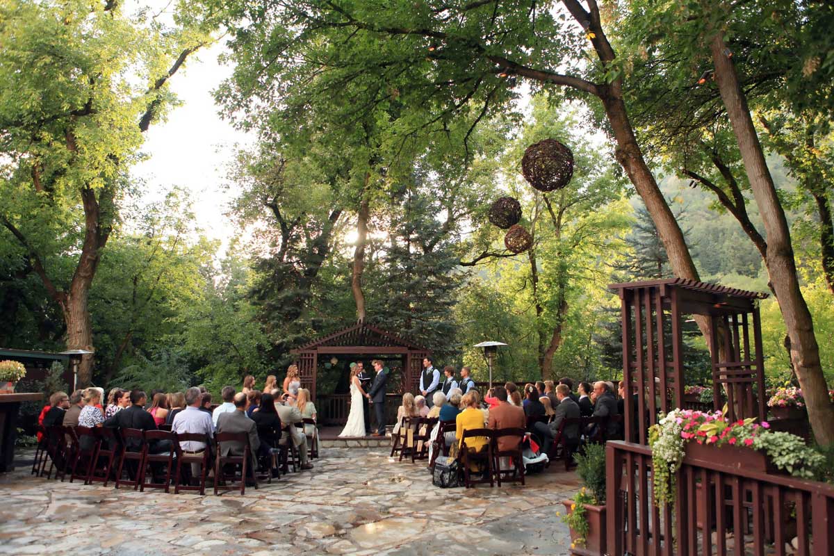 Outdoor wedding ceremony in a wooded area with guests seated on wooden chairs, a couple standing at the altar with an officiant, and decorations hanging from trees.